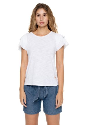 Une femme porte un T-shirt blanc à manches volantées et un short en jean bleu sur un fond blanc uni.