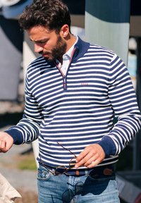 Marineblauwe en witte gestreepte kwart-rits sweater met ribgebreide manchetten en kraag, voorzien van een klein rode logo-detail en gedragen met denim jeans.
