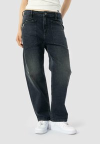 Jean en denim bleu foncé avec une coupe ample, coutures visibles et deux poches arrière. Assortis à des baskets blanches sur un fond neutre.