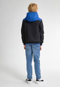 Giacca nera e blu con cappuccio, abbinata a jeans blu chiaro e sneaker bianche con dettagli neri. Vista da dietro.