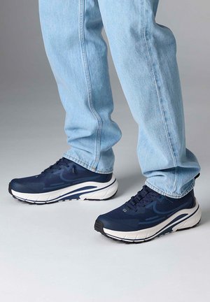 Person trägt hellblaue Jeans und dunkelblaue Sportschuhe mit weißen Sohlen und steht auf einem hellgrauen Boden.