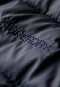 Zwarte gewatteerde jas van stof met een gestructureerde afwerking. Voorzien van een geborduurd "SUPERDRY" logo in donkere draad. Glad, duurzaam textuur.