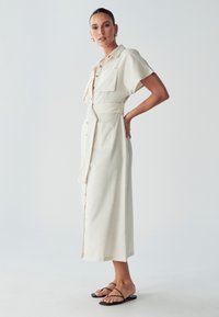 Cremefarbenes Leinenkleid mit kurzen Ärmeln, Knopfleiste, zwei Brusttaschen und einem gebundenen Gürtel in der Taille. Schwarze Sandalen mit Riemchen.
