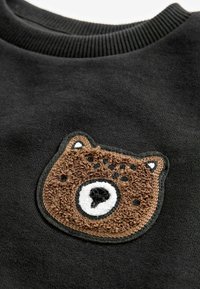 Sweat-shirt noir avec une broderie texturée représentant un visage d'ours brun avec des accents blancs et noirs au niveau de la poitrine. Texture de tissu douce.