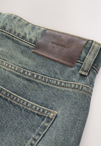 Gros plan sur un jean en denim bleu montrant la poche arrière, la boucle de ceinture et une étiquette en cuir marron avec l'inscription « DENIM SUPPLY » embossée.