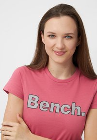 T-shirt rose à manches courtes avec le grand texte noir "Bench." et un motif à pois, fabriqué en coton doux.