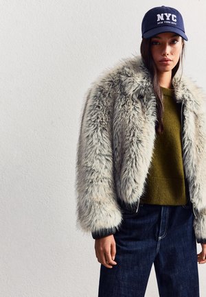 Faux fur jas in het grijs met een volumineuze textuur, gecombineerd met een donkergroene trui en donkere spijkerbroeken. Marineblauwe pet met de tekst "NYC".