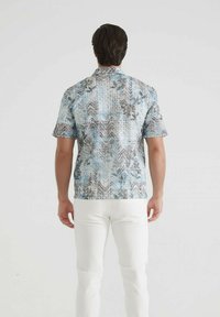 Homme se tenant de dos, portant une chemise à manches courtes bleue à motifs et un pantalon blanc, sur un fond clair et uni.