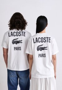 Du žmonės stovi atgal, vilkėdami baltas Lacoste Paris marškinėlius su juodais krokodilo logotipais, vieni su mėlynomis džinsomis, kiti su baltomis sijonais.