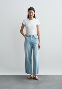 Młoda kobieta stoi z jedną ręką w kieszeni, ubrana w dopasowaną białą koszulkę, jasnoniebieskie szerokie dżinsy oraz metaliczne płaskie sandały.