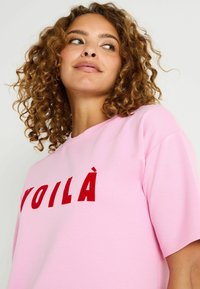 Camiseta de algodón de color rosa claro con mangas cortas y un gráfico de texto rojo que dice "VOILÀ" en el frente. Textura suave, diseño casual.