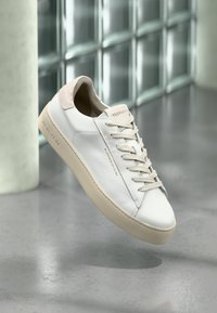 Sneaker bianco in pelle con tallone in suede beige, lacci piatti e logo impresso discreto. Presenta una suola beige chiaro e un design minimalista.