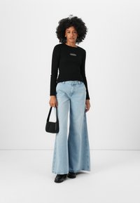 Svart langermet topp med logo, lyseblå vide jeans og svart håndveske. Modellen har på seg svarte lakk sko; enkel hvit bakgrunn.