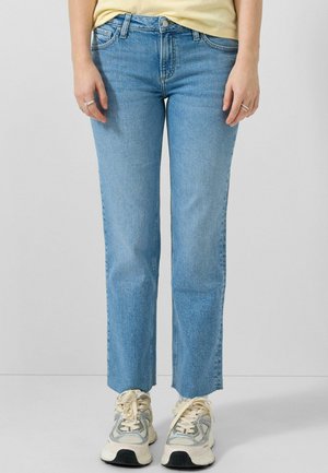 Osoba nosząca niebieskie dżinsy o prostych nogawkach, zestawione z kremowo-szarymi trampkami, stojąca na tle jednolitego jasnoszarego tła.