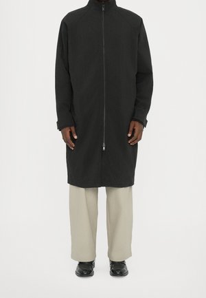 Homme portant un long manteau noir zippé, un pantalon beige clair et des chaussures noires, debout devant un fond blanc.