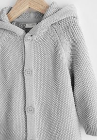 Cardigan bébé en tricot gris clair avec capuche, trois boutons à l’avant et motifs texturés, suspendu sur un cintre en bois sur fond blanc.