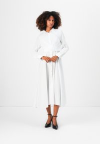 Abito bianco a maniche lunghe con chiusura a bottoni frontale, design avvolgente in vita e orlo fluente. Abbinato con scarpe nere con tacco alto.