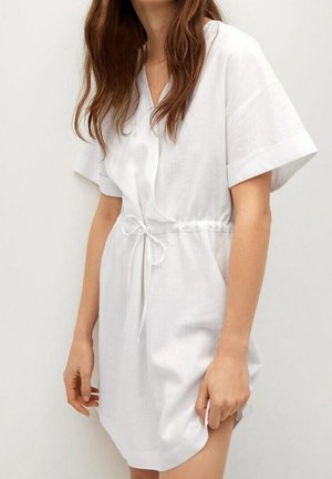 Kobieta w białej sukience z krótkim rękawem i paskiem w talii stojąca na tle jasnego, jednolitego tła.