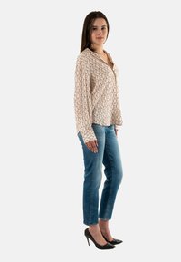 Blusa de manga larga en beige claro con un patrón geométrico, combinada con jeans azul claro y tacones altos negros, de pie frente a un fondo liso.