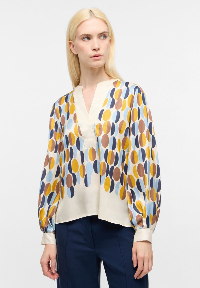 Blusa a maniche lunghe in tessuto crema con un motivo circolare colorato in blu, giallo, marrone e blu navy. Presenta una scollatura a V e maniche arricciate.