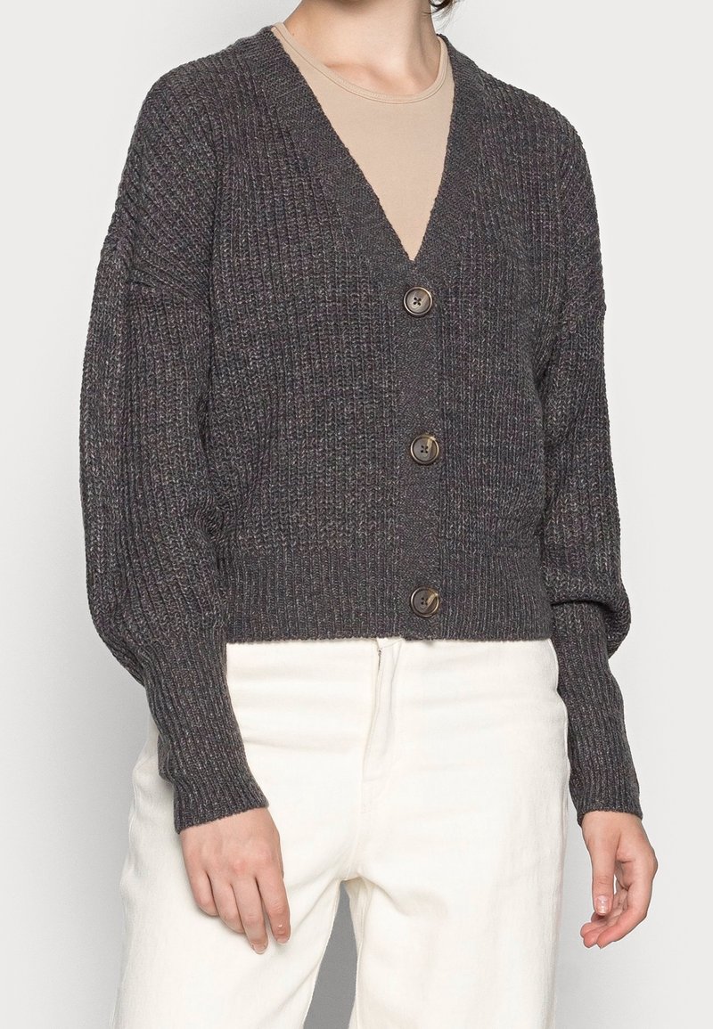 Personne portant un cardigan en maille gris foncé à trois boutons sur un haut beige et un pantalon blanc sur un fond uni.