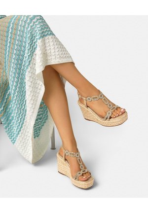Beine überschlagen, beige Keilsandalen mit zirkulären Perlendetails und ein blau-weiß-tan gestreiftes, zickzackgemustertes Strickkleid tragend.