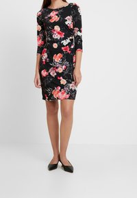 Zwarte bloemenjurk met driekwart mouwen, voorzien van roze en witte bloemenpatronen. Glad stof met een aansluitende silhouet, gecombineerd met zwarte schoenen.