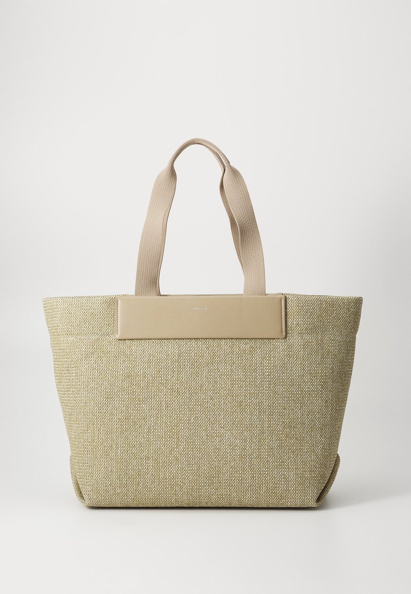 Sac fourre-tout tissé beige avec des anses larges en tissu et un panneau en cuir lisse près du haut, sur un fond blanc uni.