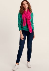 Groene gebreide trui, roze sjaal, donkerblauwe spijkerbroek en witte sneakers. De sjaal heeft een gestructureerd golvend patroon, terwijl de spijkerbroek slim fit is.