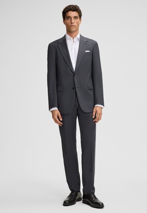 Costume gris foncé en tissu texturé. Comprend une veste à boutonnière simple, des revers en pointe et un pantalon assorti, associé à une chemise habillée blanche.