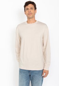 Maglietta beige a maniche lunghe in morbido cotone, collo tondo, con un logo sottile sul davanti, abbinata a jeans in denim blu chiaro.