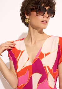 Sukienka z dekoltem w serek w kolorowy abstrakcyjny wzór w odcieniach różu, pomarańczowego i kremowego, wykonana z gładkiego materiału. Złote kolczyki w kształcie kwiatów i duże okulary przeciwsłoneczne.