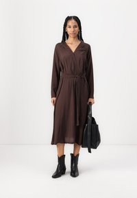Robe marron à manches longues, ceinturée à la taille et avec une jupe fluide. Portée avec des bottines noires et tenant un sac noir. Texture de tissu lisse.