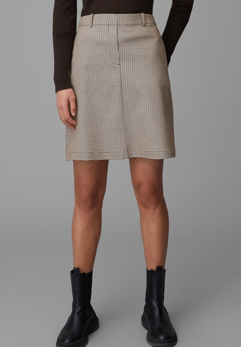 Femme portant une jupe beige et noire à carreaux jusqu'aux genoux, un haut noir à manches longues et des bottines noires sur un fond gris uni.