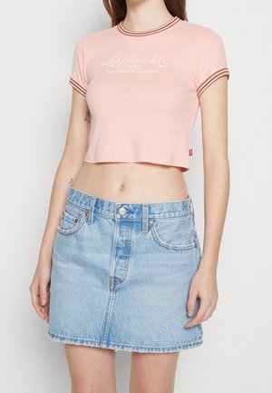 Kobieta nosząca różowy krótki t-shirt z czerwonym wykończeniem i logo, zestawiony z jasnoniebieską mini spódnicą jeansową z wysokim stanem.