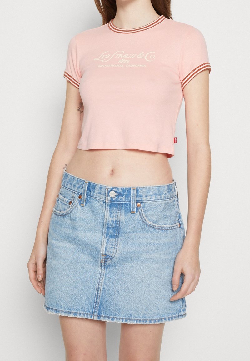 Femme portant un t-shirt court rose avec des bordures rouges et un logo, associé à une mini-jupe en denim bleu clair taille haute.
