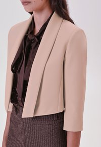 Beige cropped blazer met een gladde textuur, met notch revers en driekwart mouwen, gedragen over een donkerblouse en pantalon met pied-de-poule patroon.