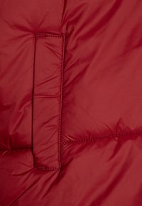 Red puffy jacket fabric with a smooth texture, featuring a rectangular pocket and visible stitching along quilted sections.