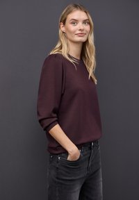 Młoda kobieta z długimi blond włosami, ubrana w bordową bluzkę z długim rękawem i czarne dżinsy, stojąca z jedną ręką w kieszeni na ciemnym tle.