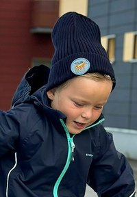 Mörkblå ribbad stickad mössa med en logopatch och uppvikt kant, bärs med en mörk jacka med en ljus turkos dragkedja och detaljer.