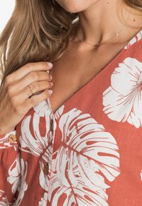 Bluzka w kolorze rdzy z białym wzorem liści, dekoltem w serek i drewnianymi guzikami. Posiada teksturowany materiał oraz delikatny złoty naszyjnik z koralikami.