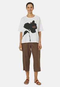 Witte t-shirt met een groot zwart bloemenpatroon, gecombineerd met losse bruine linnen capri-broeken en bruine sandalen. Casual outfit met een relaxte pasvorm.