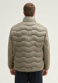 Puchowa kurtka w jasnym oliwkowym kolorze z falistą, pikowaną teksturą. Posiada wysoki kołnierz i elastyczne mankiety, gładkie boczne panele oraz minimalistyczne detale.