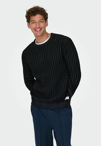 Crni zdravi pulover s okomitim plavim prugama, okruglim izrezom i rebrastim manžetama. Uparen s mornarskim hlačama i bijelom majicom.