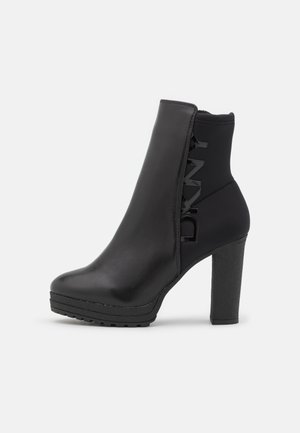 Bottine noire à talon haut avec talon épais, semelle crantée, avant en cuir lisse et détail brillant du logo de la marque sur la tige latérale.