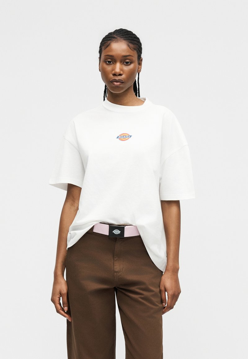 Giovane donna con capelli intrecciati che indossa una camicia bianca a maniche corte con un logo e pantaloni marroni con una cintura rosa chiaro.