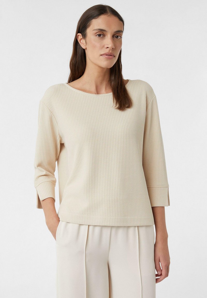 Top lungo a maniche ribatte con scollatura a barca e maniche con risvolto, abbinato a pantaloni a gamba larga beige chiaro. Tessuto morbido e testurizzato.