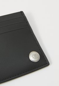 Portefeuille en cuir noir avec plusieurs emplacements pour cartes, accent en logo circulaire argenté et bords cousus, affichant une texture lisse.