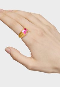 Złoty pierścionek z różowym kamieniem szlachetnym w kształcie kwiatu, o gładkiej, błyszczącej powierzchni i zaokrąglonych krawędziach, noszony na dłoni.