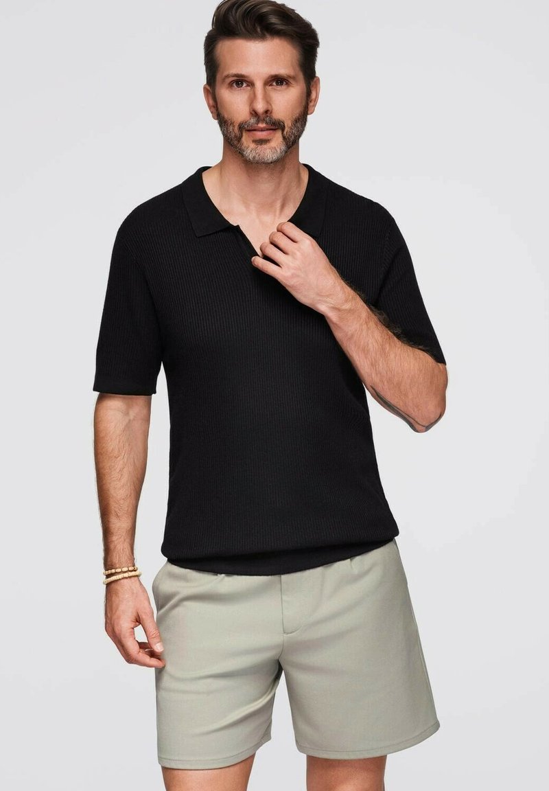 Černé pruhované polo tričko s plochým límcem, spárované se světlými šedými šortkami. Model má vousy a je v uvolněné pozici.